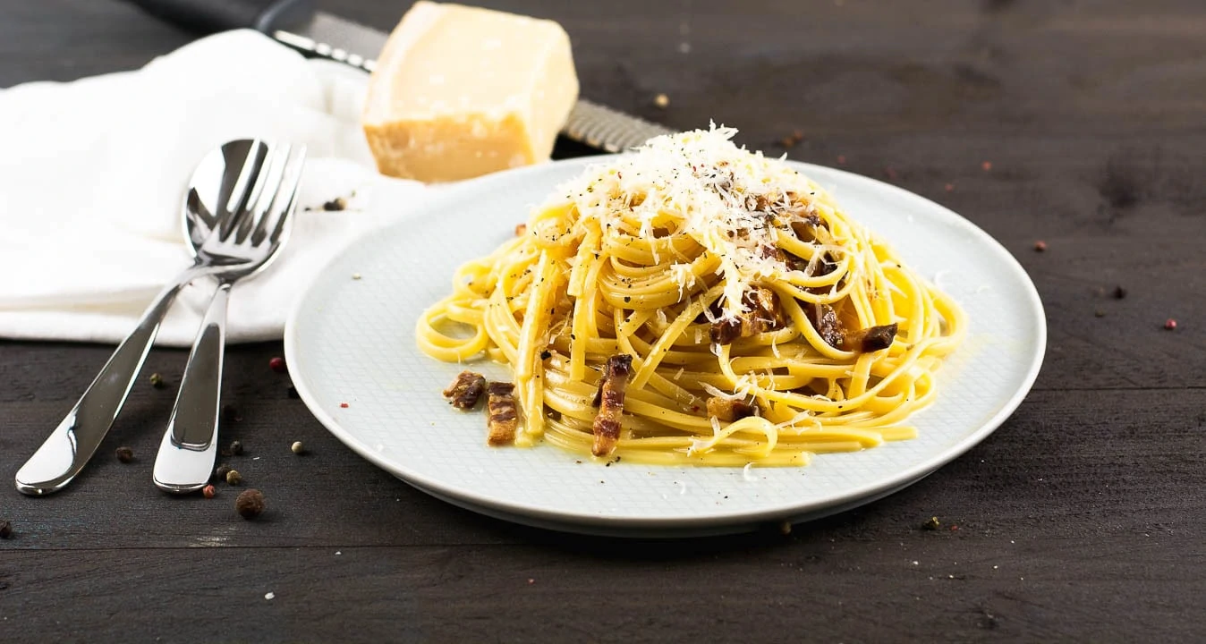 Originales Spaghetti Carbonara Rezept ohne Sahne mit Parmesan bedeckt auf einem Teller. Im Hintergrund ist ein Stück Parmesan und eine Reibe.