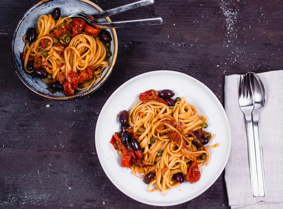 Zwei Teller Spaghetti Puttanesca von oben auf einem schwarzen Tisch
