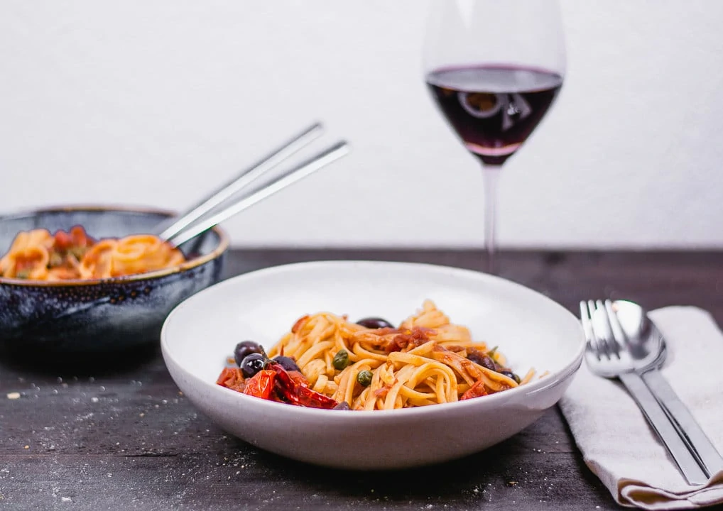 Ein Teller Spaghetti Puttanesca mit einem Glas Rotwein im Hintergrund