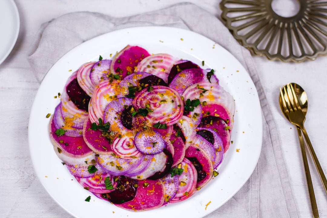 Eine goldene Gabel und ein goldener Löffel neben einem Teller bunte Bete Carpaccio.