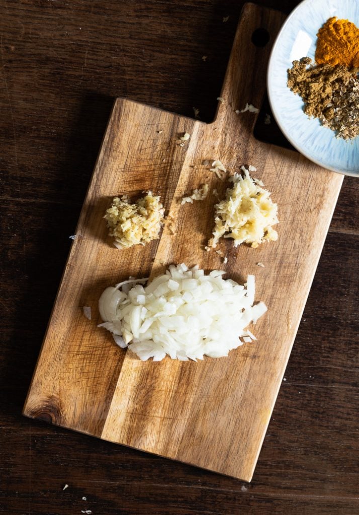 Zwiebeln, Ingwer und Knoblauch liegen geschnitten auf einem Brett für das Kichererbsen Gericht.