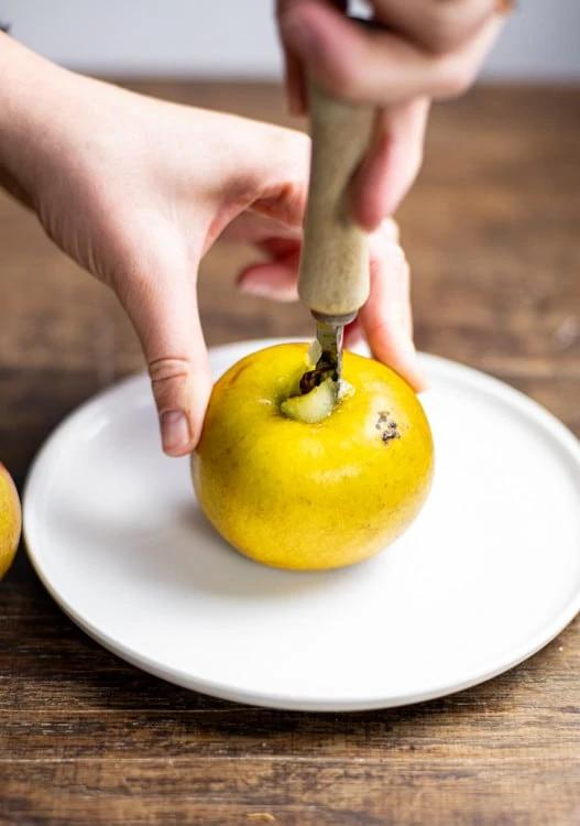 Ein Boskop Apfel wird für das Bratapfel Rezept mit Hilfe eines Apfelentkerners entkernt. Der Apfel steht dafür auf einem weißen Teller. 