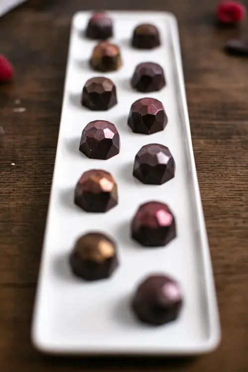 Himbeer Pralinen in Diamantenform liegen auf einem länglichen Teller. Sie glänzen leicht rosa.