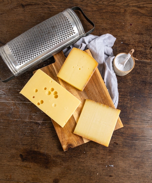 Eine Auswahl von drei Käsen liegt auf einem Brett auf dem Tisch. Zu sehen sind Appenzeller, Le Gruyere und Emmentaler. Außerdem mit im Bild ist eine Reibe und ein Schälchen mit Salz. 
