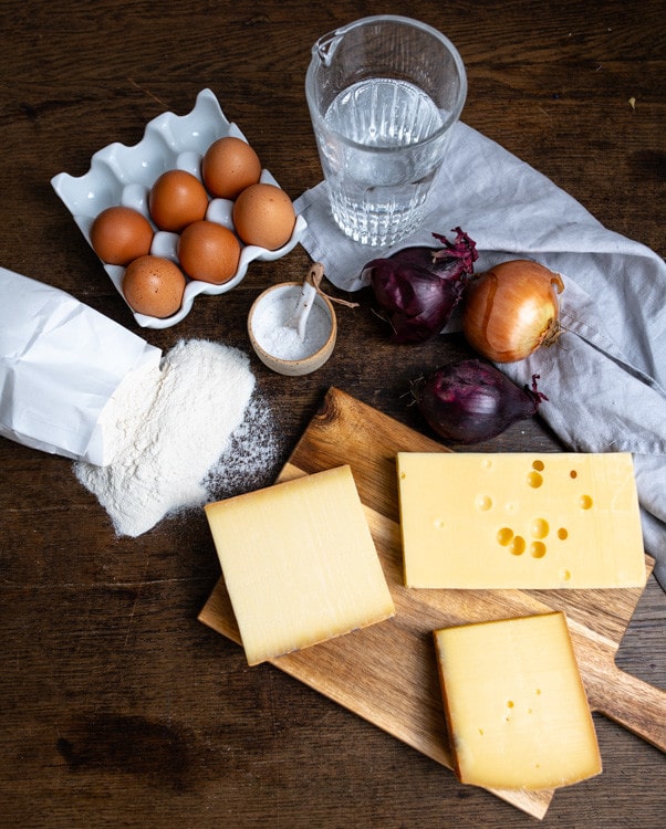 Die Zutaten, die für die Käsespätzle benötigt werden liegen auf dem Tisch. Im Uhrzeigersinn sind das Eier, Salz, Mineralwasser, Zwiebeln, verschiedene Bergkäse und Mehl.