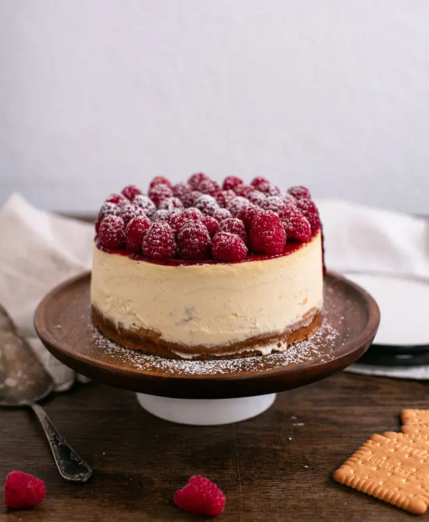 Ein ganzer New York Cheesecake mit Himbeeren steht auf einer Kuchenplatte aus Holz. Diese steht etwas erhöhe. Auf dem Tisch um den Kuchen herum liegen Butterkekse und Himbeeren. Die Himbeeren auf den Cheesecake mit sind mit etwas Puderzucker bestreut.