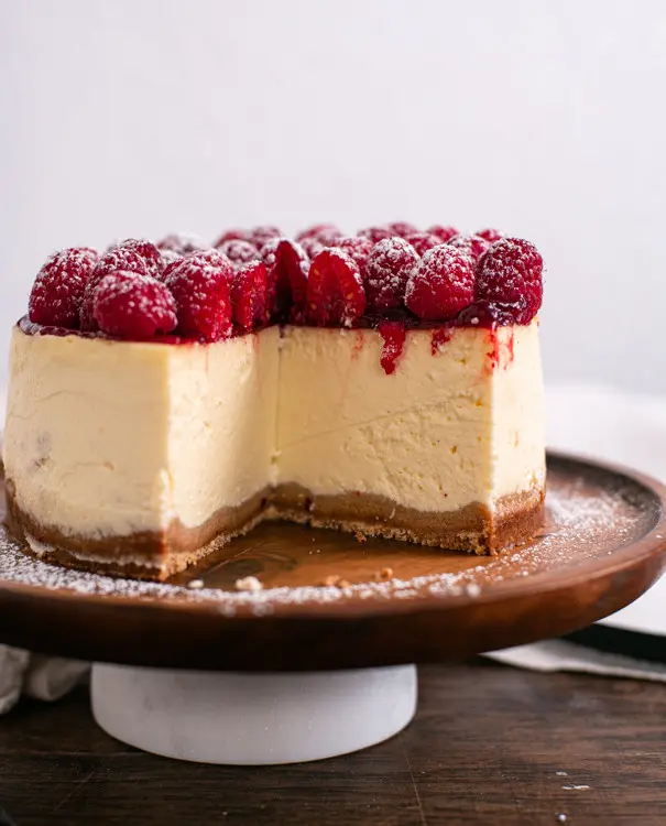 Auf diesem Bild ist der New York Cheesecake angeschnitten. Es fehlt etwa ein viertel. Im Anschnitt erkennt man die kompakte, cremige Textur der Cheesecake Creme. Der New York Cheesecake ist oben mit reichlich frischen Himbeeren dekoriert.