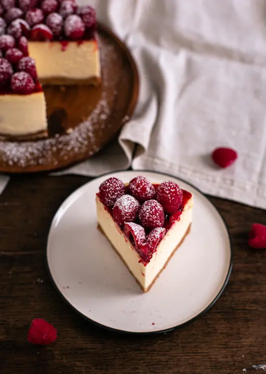 Ein Stück des New York Cheesecake steht auf einem kleinen Teller. Das Foto ist von oben Fotografieren. Auf den Himbeeren befindet sich etwas Puderzucker. Im Hintergrund ist der Rest des Cheesecake zu sehen.