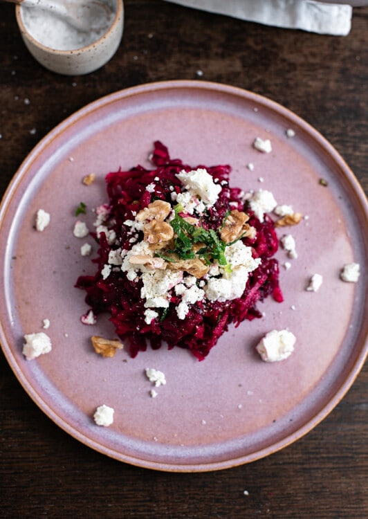 Rote Beete Salat mit Feta, Walnüssen und Minze auf einem rosa Teller angerichtet. Im Hintergrund ist ein Schälchen mit Salz zu sehen. 