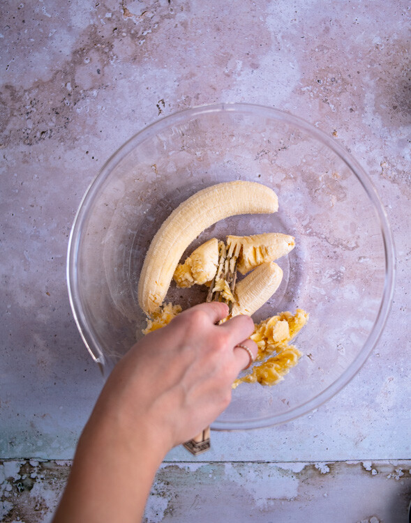 Drei große, reife Bananen werden für das vegane Bananenbrot in einer großen Schüssel mit einer Gabel zerdrückt. 