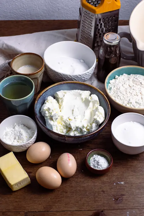 Die Zutaten für das Käsekuchen Rezept liegen auf dem Tisch. Das sind, Milch, Zucker, Vanileextrakt, Öl, Quark, Mehl, Stärke, Zucker, Eier, Butter und Backpulver