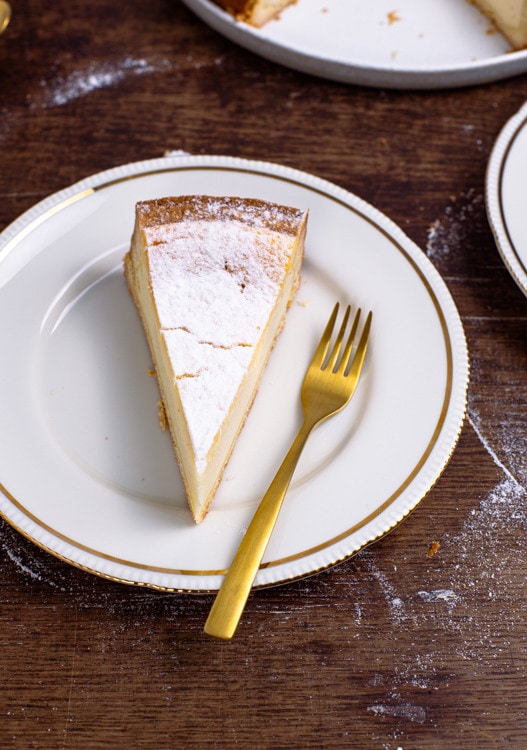 Ein Stück Käsekuchen liegt auf einem weißen Teller mit Goldrand. Eine goldene Gabel liegt neben dem Kuchenstück auf dem Teller. 