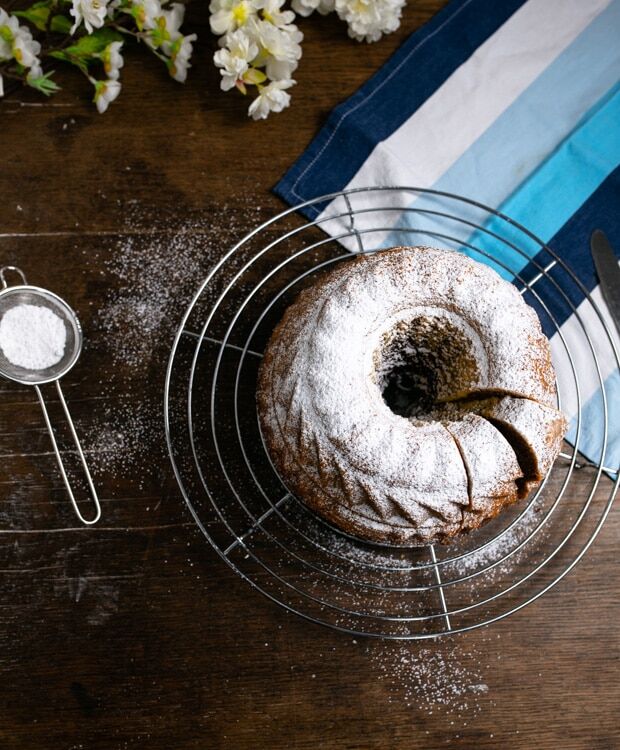 Marmorkuchen, gebacken in einer Guglhupfform steht auf einem Kuchengitter. Der Kuchen ist mit Puderzucke bestreut und es sind zwei Stücke vom Kuchen abgeschnitten. 