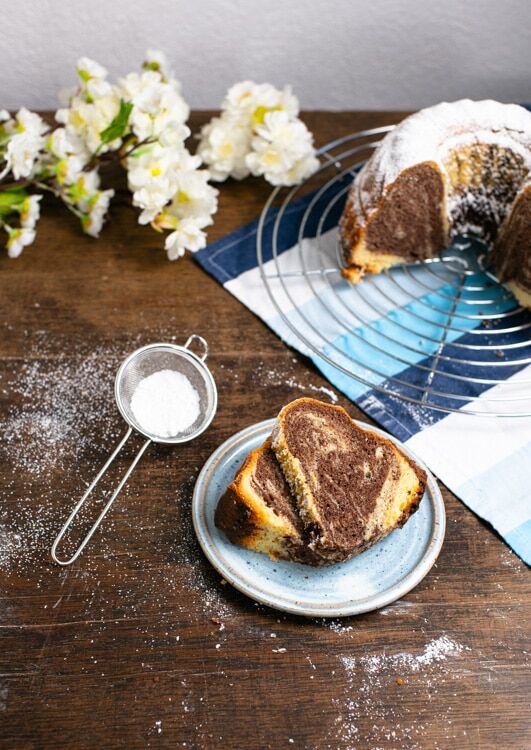 Zwei Stücke Marmorkuchen liegen auf einem hellblauen, handgetöpferten Teller. Das Marmormusterals hellem und dunklem Teig ist gut zu erkennen. Neben dem Teller liegt ein Sieb mit Puderzucker. Der restliche Kuchen steht auf einem Kuchengitter.