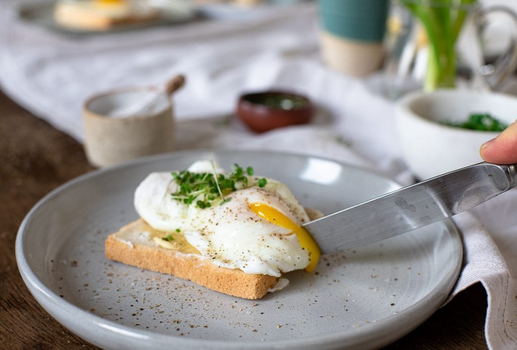 Zwei pochierte Eier liegen auf einer Scheibe Toastbrot. Eines der Eier wird mit einem alten, silbernen Messer angeschnitten. Das Eigelb fließt heraus. 