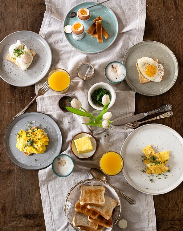 Fünf verschiedene Frühstückseier stehen auf einem gedeckten Brunch Tisch. Es gibt weich gekochte Eier, pochierte Eier, Spiegeleier, Rührei und Omelette. 