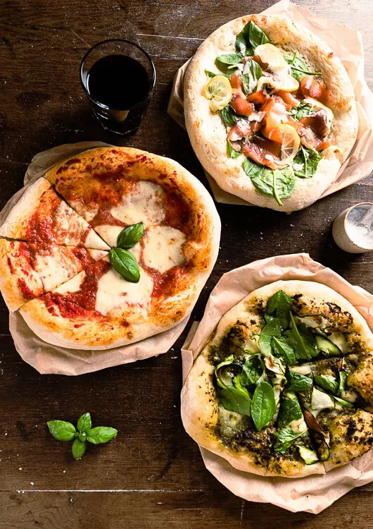 Eine Pizza Margarita, eine Pizza mit Pesto und eine Pizza mit Sauerrahm und Räucherlachs liegen fertig gebacken auf dem Tisch. 