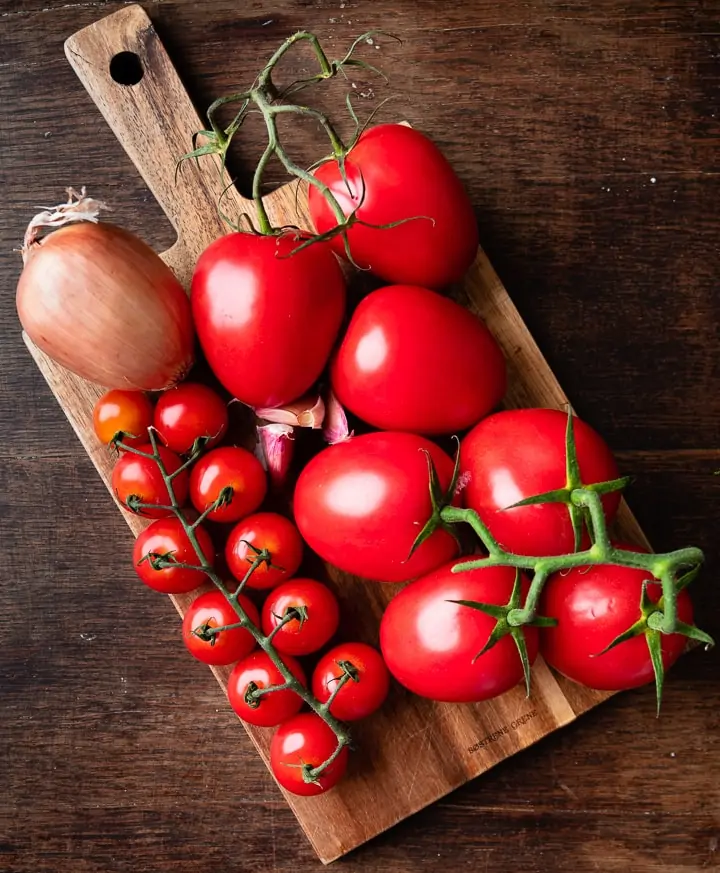 Mehrere verschiedene Tomaten, zwiebeln und Knoblauch liegen auf einem Holzbrett.