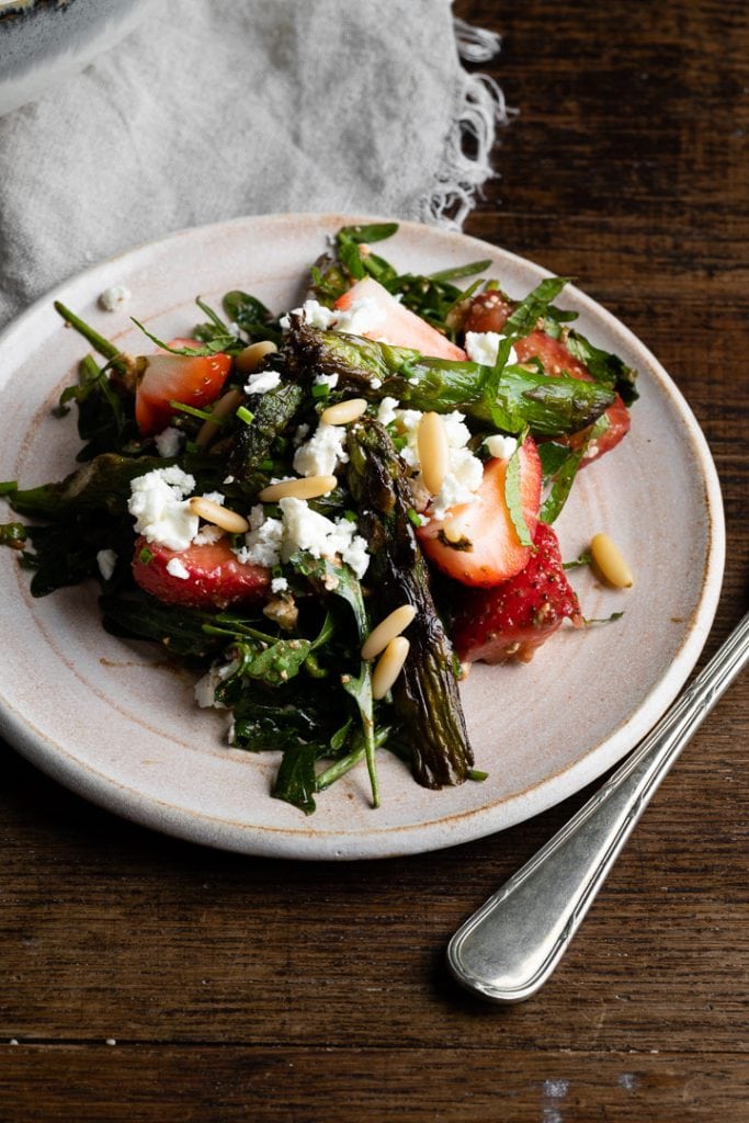 Salat aus gebratenem grünen Spargel, frischen Erdbeerscheiben, zerbröckeltem Feta-Käse und Pinienkernen auf einem Bett aus Blattsalat, serviert auf einem hellen Teller.