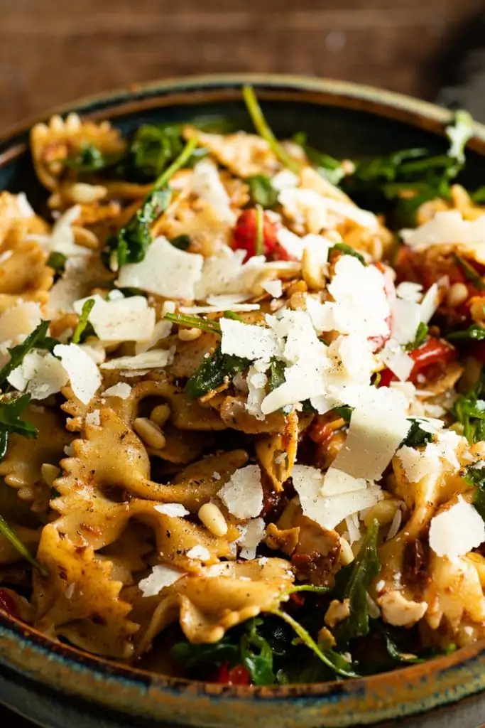 Mediterraner Farfalle-Salat mit Rucola, getrockneten Tomaten und Pinienkernen, großzügig bestreut mit Parmesan-Spänen, in einer dunklen Schale.
