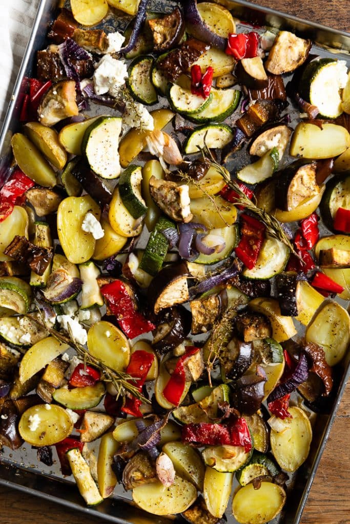 Geröstetes Ofengemüse auf einem Backblech: Kartoffelspalten, Zucchini, Paprika, rote Zwiebeln und eventuell Aubergine, gemischt mit Feta-Käse und Kräutern wie Thymian und Rosmarin.
