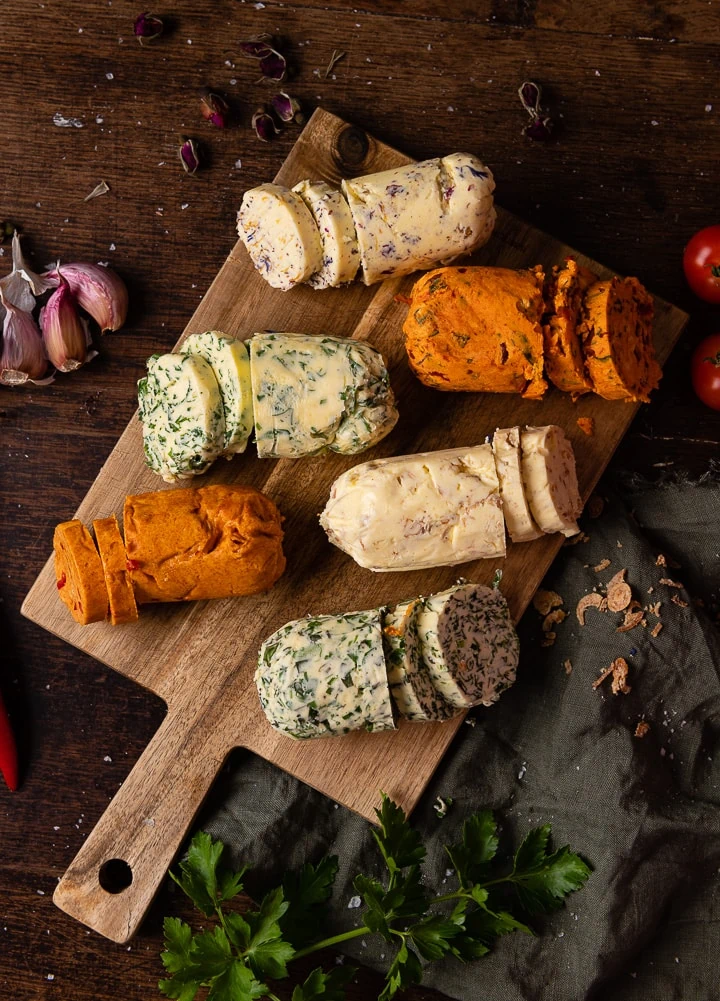 Sechs Rollen selbstgemachter Kräuterbutter und Tomatenbutter, aufgeschnitten auf einem Holzbrett mit Kräutern und Knoblauch dekoriert.
