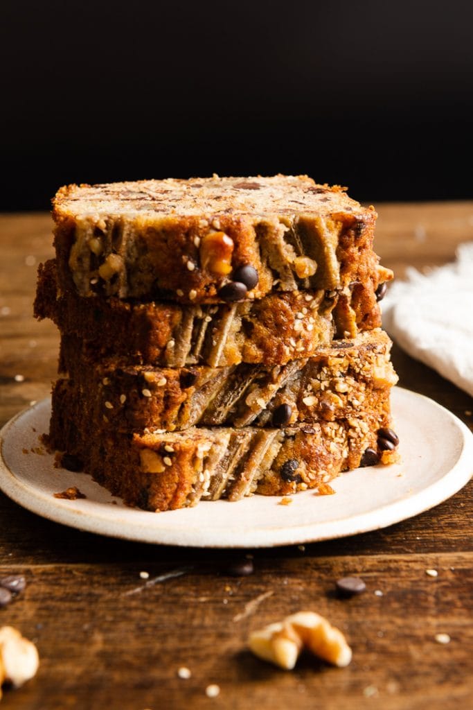 Vier dicke Scheiben Bananenbrot mit Walnüssen und Schokostückchen, auf einem hellen Teller gestapelt, auf einem Holztisch.