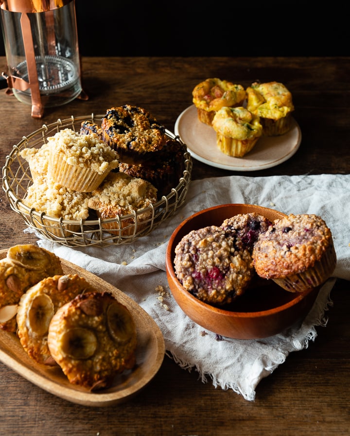 Auswahl an Frühstücksmuffins auf einem Holztisch: Streuselmuffins im Korb, herzhafte Eimuffins auf Teller, Hafer-Bananen-Muffins auf Holzbrett und Beeren-Hafermuffins in Schale.