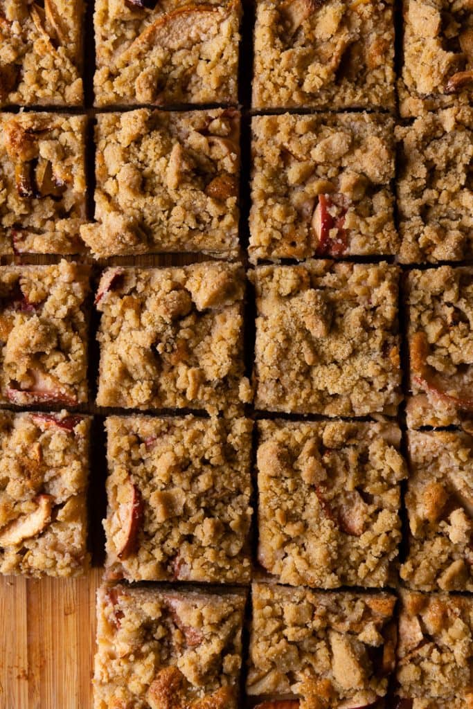 Quadratische Stücke von Apfel-Streuselkuchen vom Blech, auf einem Holztisch angerichtet. Man sieht Apfelstückchen unter der Streuseldecke.