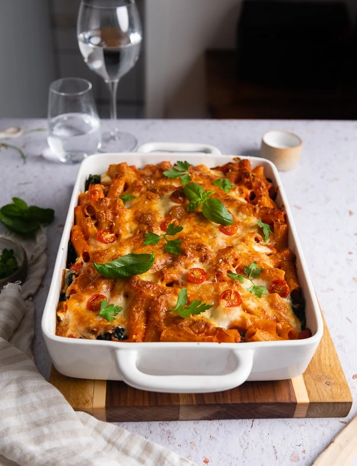 Vegetarischer Nudelauflauf mit Rigatoni (oder ähnlicher Pasta) und Spinat, goldbraun mit Käse überbacken, serviert in einer weißen Auflaufform auf einem Holzbrett.