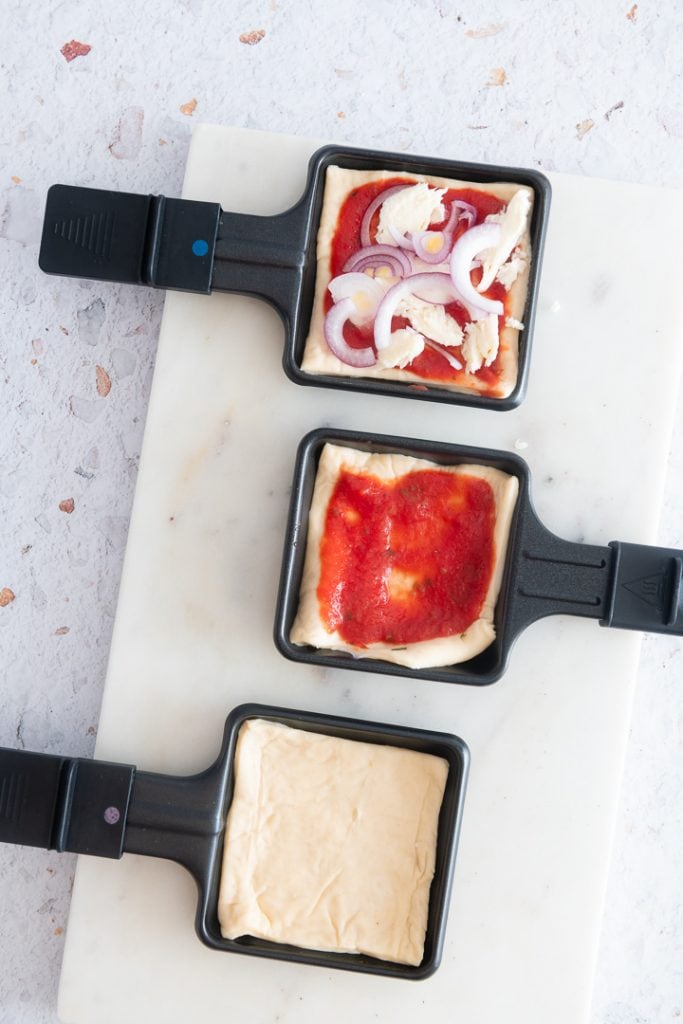 Drei Raclettepfännchen sind mit Pizzateig, Tomatensauce und Mozzarella und Zwiebeln befüllt.