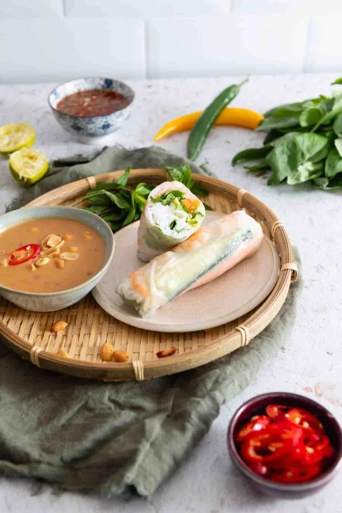 Zwei vegetarische vietnamesische Sommerrollen aus Reispapier mit Gemüsefüllung, serviert auf einem Bambustablett mit Erdnuss-Dip und weiterem Dip.
