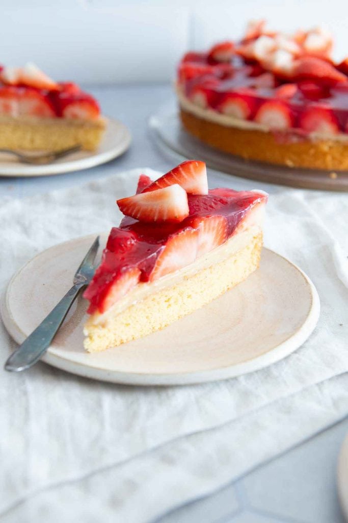 Ein Stück Erdbeerkuchen mit Biskuitboden & Vanillepudding, daneben liegt eine Gabel.
