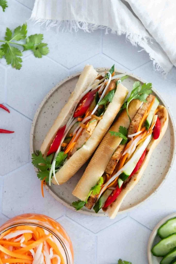 Zwei Sandwiches auf einem Teller in der Tischmitte. Daneben ist das eingelegte asiatische Gemüse.