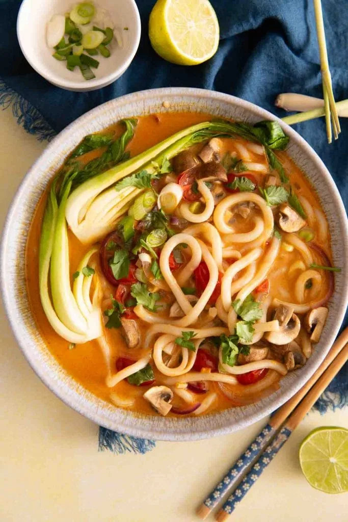Eine Schüssel Thai-Curry-Suppe mit dicken Udon-Nudeln, Pak Choi, Champignons, Paprika und roten Zwiebeln in einer Kokos-Curry-Brühe, garniert mit Koriander und Frühlingszwiebeln.