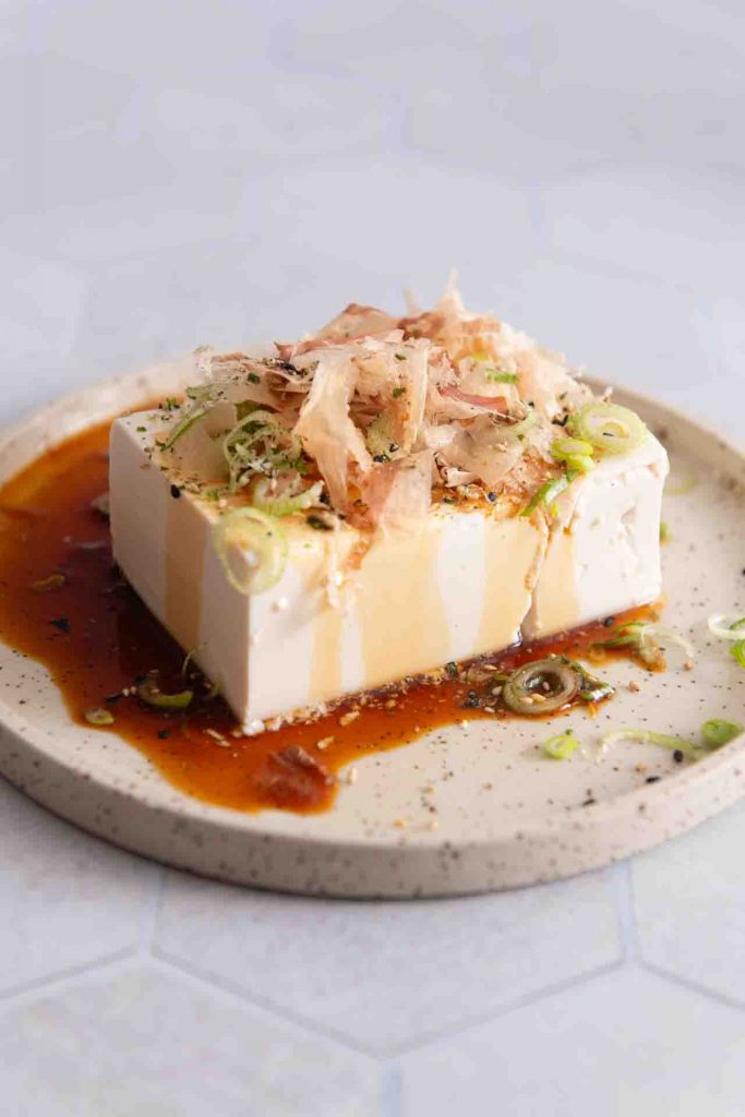 Ein Block Seidentofu serviert als Hiyayakko auf einem kleinen Teller, garniert mit Katsuobushi (Bonitoflocken), gehackten Frühlingszwiebeln und einem Schuss Sojasoße.