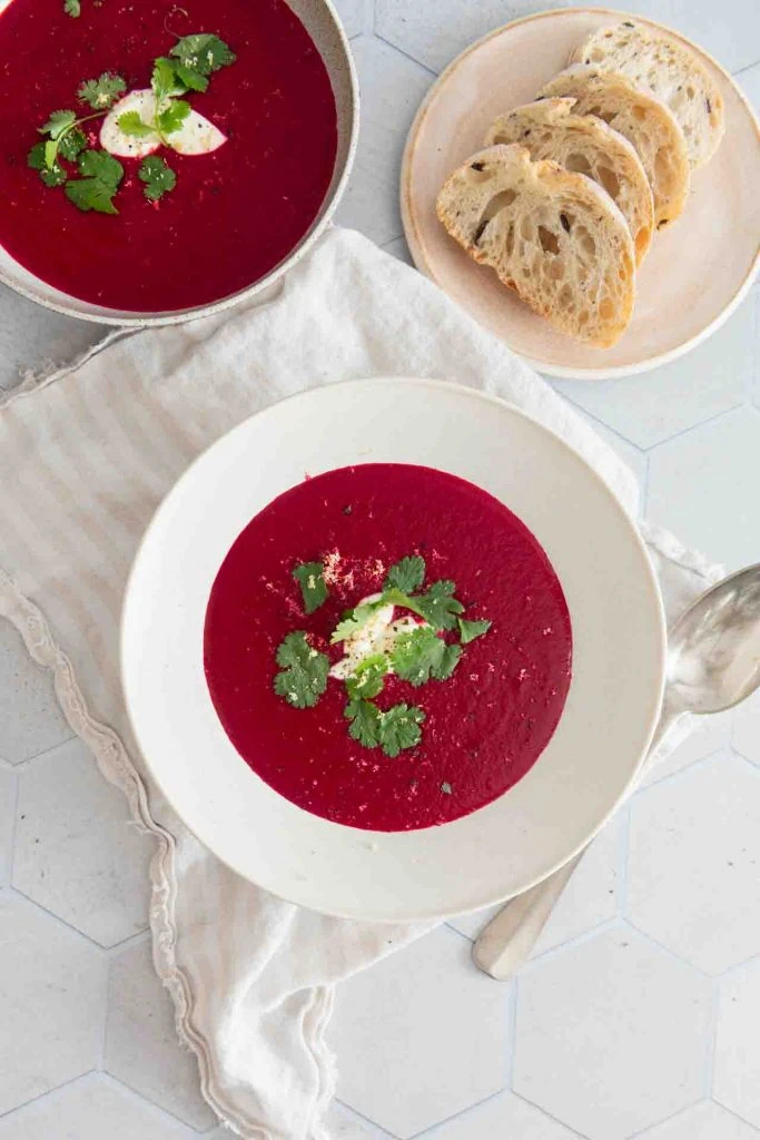 Zwei Teller mit Rote Bete Suppe mit Kokosmilch stehen auf einem gedeckten Tisch, zu sehen sind auch einige Scheiben Weißbrot, die mit der Suppe serviert werden.