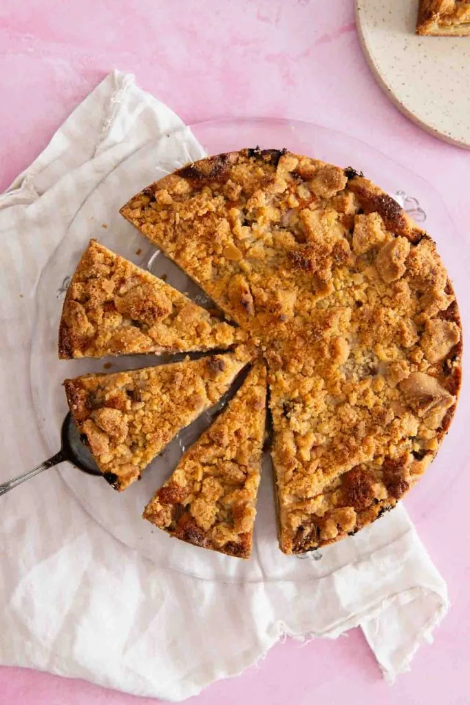 Der Rhabarberkuchen mit Streuseln liegt auf einer Kuchenplatte aus Glas. Er ist von oben fotografiert und es sind vier Stücke des Kuchens angeschnitten. Unter einem der Stücke liegt eine Tortenschaufel. Ein Stück fehlt und ist am oberen rechten Rand zu erkennen.
