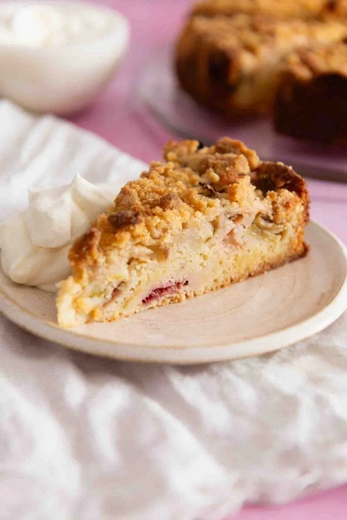 Ein Stuck Rhabarberkuchen mit Streuseln liegt auf einem Teller. Auf dem Teller befindet sich außerdem ein Klecks Schlagsahne. Im Hintergrund ist der restliche Kuchen zu sehen.