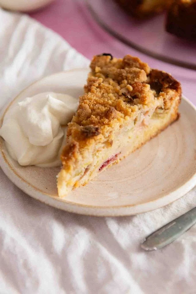 Ein Stück Rhabarberkuchen mit Streuseln liegt auf einem Kuchenteller mit einem Klecks Schlagsahne.