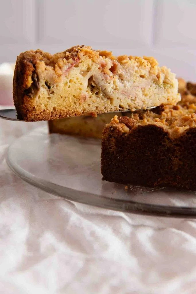 Ein Stück des Rhabarber Streuselkuchens wird mit einer Kuchenschaufel von der Kuchenplatte gehoben.